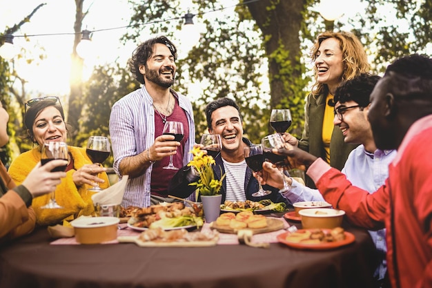 Group of friends dining outdoors toasting with red wine in the backyard multi generational diverse people having fun together in the nature sitting around the dinner table focus on the mature man