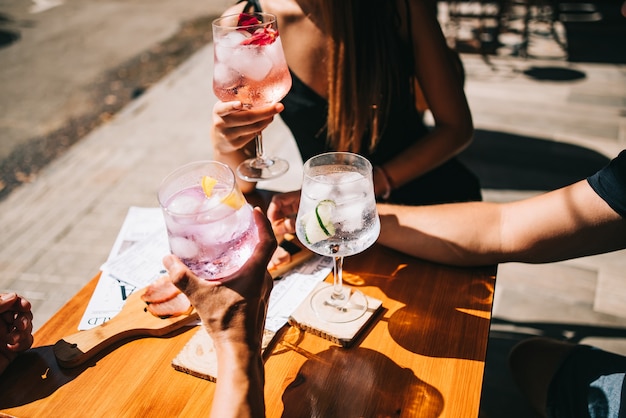 Group of friends sitting on the summer terrace holding cocktails and snacks on the table