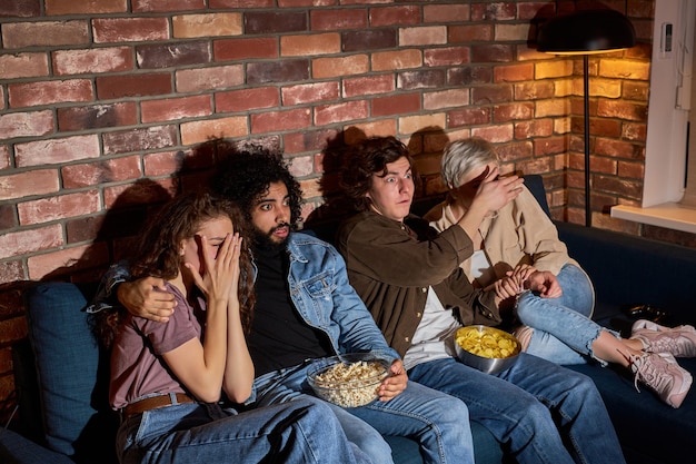 Group of friends watching scary movie on TV at home, eating popcorn, sitting on sofa together, closing eyes, two couples nervous scared in shock terrified