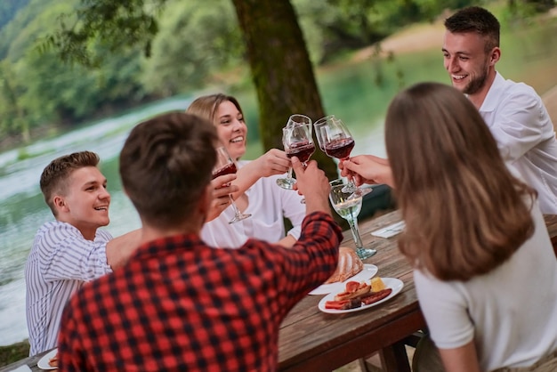 group of happy friends toasting red wine glass while having picnic french dinner party outdoor during summer holiday vacation near the river at beautiful nature