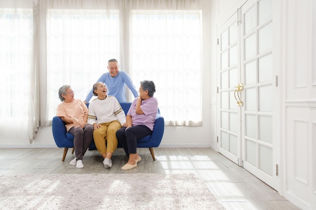 The group of happy and healthy three Asian senior women sits together on a sofa with an Asian senior man standing in the back Smiling and laughing together Image with copy space