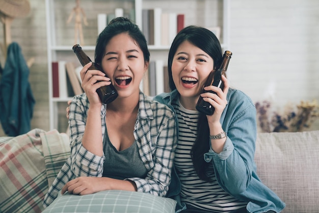 Group happy young female people sitting on sofa with bottled drinks. two asian girl best friends holding beers putting by cheeks face camera laughing smiling attractive sitting on couch at home.