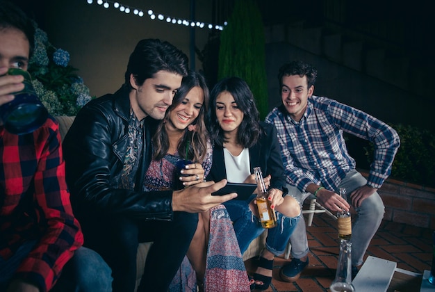 Group of happy young friends drinking and laughing while looking a smartphone picture in a outdoors party. Friendship and celebrations concept.