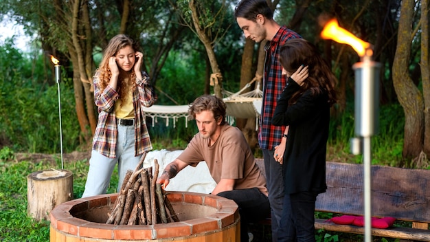 A group of happy young friends trying to make a campfire at glamping. Two men and women