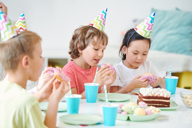 Group of hungry little kids eating glazed donuts with sprinkles and having drinks while sitting by birthday table