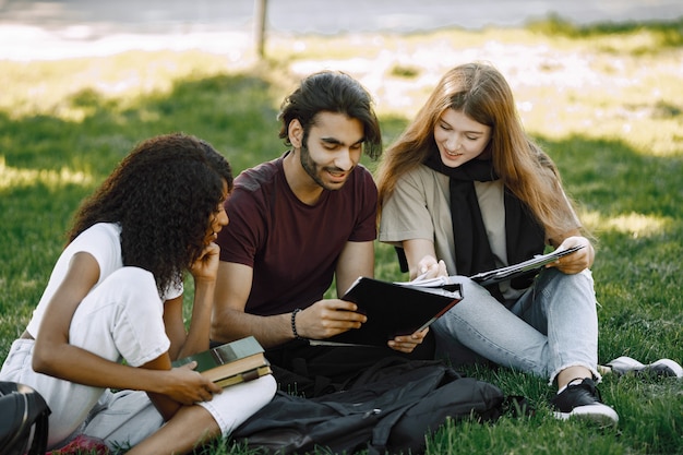 Group of international students sitting on a grass together in park at university. African and caucasian girls and indian boy talking outdoors