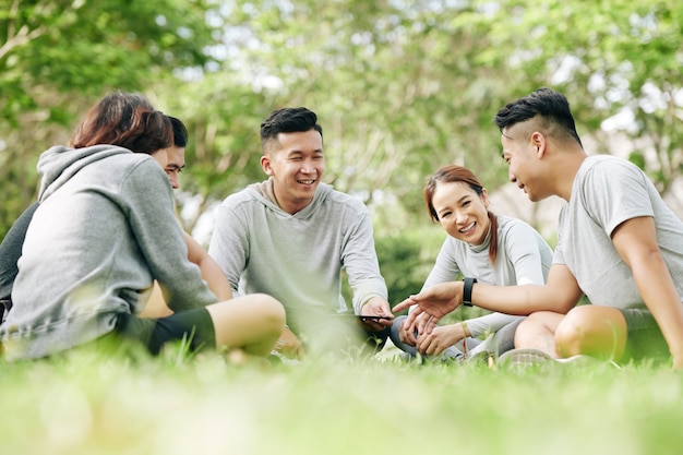 Group of joyful young students sitting on grass loan on campus and discussing new smartphone model or mobile application