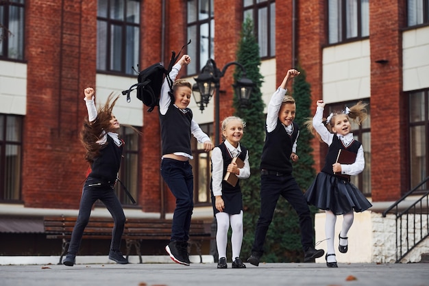 Group of kids in school uniform jumping and having fun outdoors together near education building.