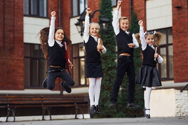 Group of kids in school uniform jumping and having fun outdoors together near education building.