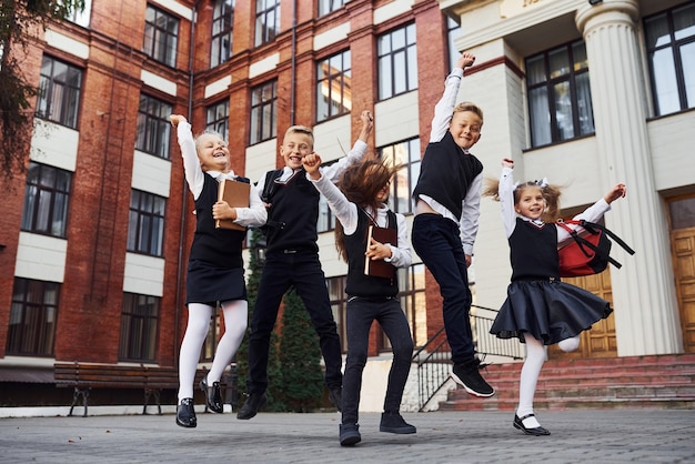 Group of kids in school uniform jumping and having fun outdoors together near education building.