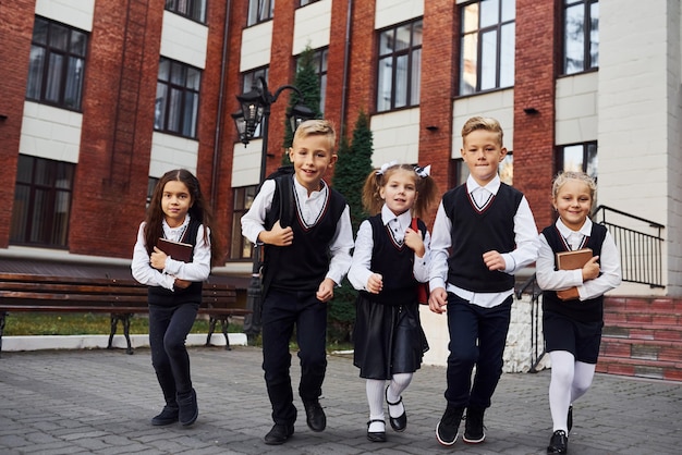 Group of kids in school uniform posing to the camera outdoors together near education building.