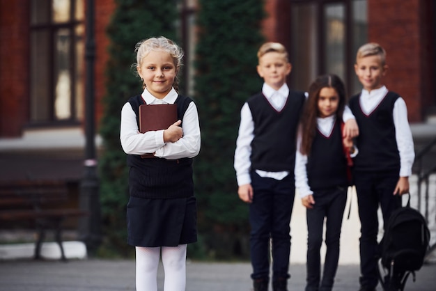 Group of kids in school uniform posing to the camera outdoors together near education building.