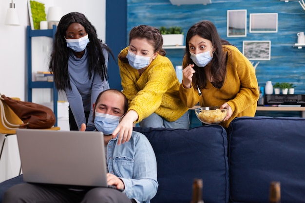 Group of mixed race friends having fun keeping social distancing with face mask preventing coronavirus spreading looking shocked at video on laptop in living room drinking beer and eating chips. Conce