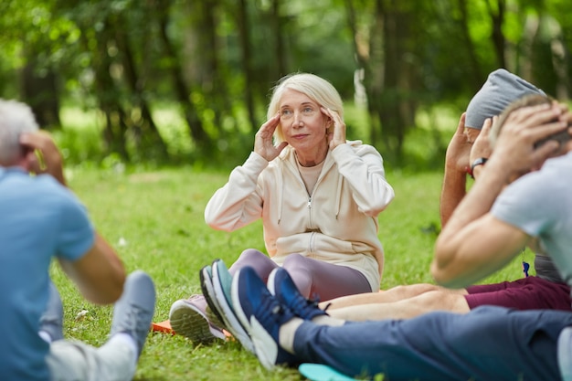 Group of modern senior people gathered together in park sitting on mats on grass doing neck stretching relaxing exercise