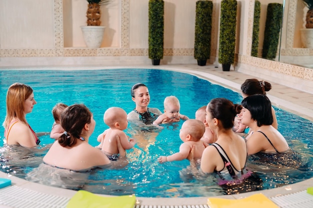 A group of mothers with their young children in a children's swimming class with a coach.