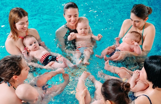 A group of mothers with their young children in a children's swimming class with a coach.