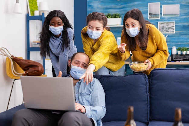 Group of multiethnic friends laughing together watching funny video on laptop in home living room wearing face mask as safety precaution, not to sprea coronavirus during global pandemic.