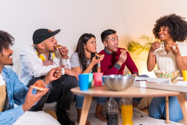 Group of multiethnic friends on a sofa eating pizza and drinking soda at a home party eating pizza and telling jokes