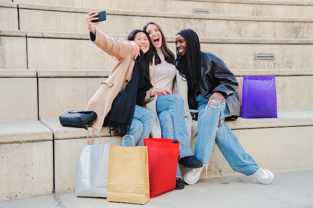 Group of multiracial young women smiling doing a selfie portrait with cellphone after shopping Three multiehnic happy girls having fun taking pictures sitting outdoors Lifestyle concept