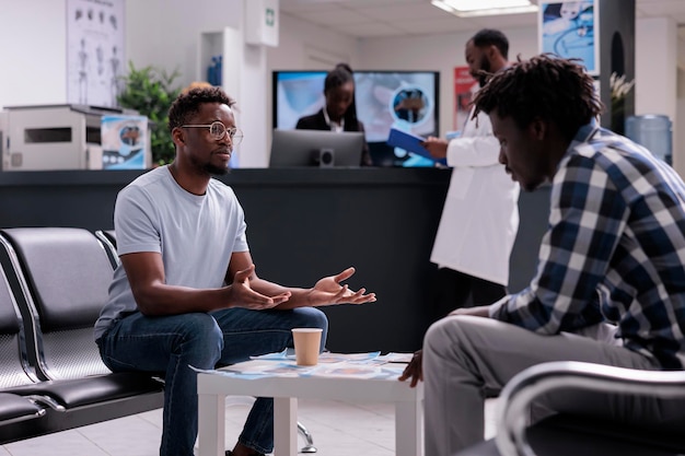 Group of patients talking in waiting area at hospital lobby before medical consultation appointment. Men sitting in waiting room at healthcare facility having conversation, checkup visit support.