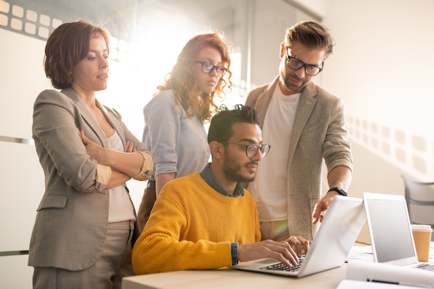 Group of pensive creative advertisers standing at desk and asking web designer to add design elements for presentation in office