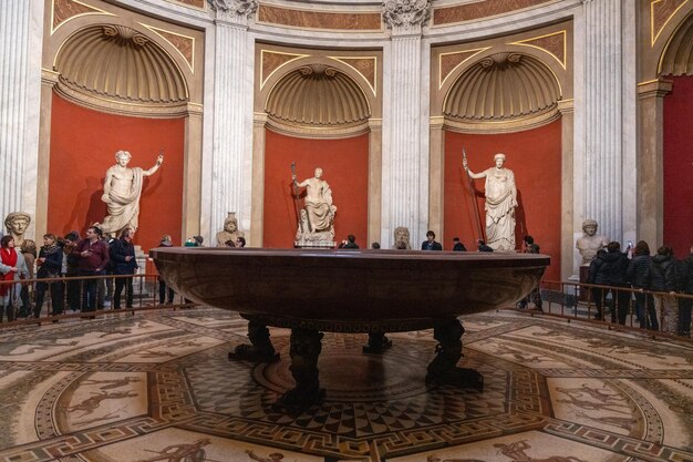 A group of people are standing around a large table in a room with statues of pe