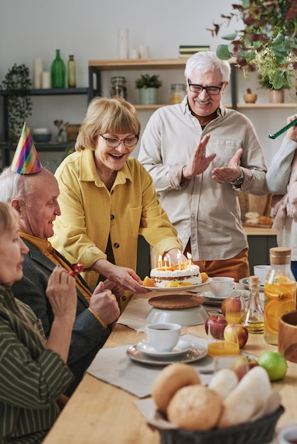 Group of senior friends celebrating birthday with cake with candles while sitting at dining table together at home