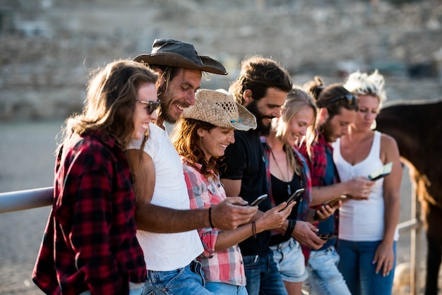 Group of seven people using their phonnes together smiling and having fun in a ranch with horses at the background - caucasasian group of adults enjoying with the technology