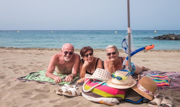 Group of smiling caucasian seniors lying on the sea beach sunbathing enjoying leisure time happy mature women and man looking at camera