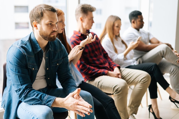 Group of stressed young diverse multi-ethnic job candidates in casual clothes waiting interview with hr, sitting in queue line row on chairs in modern office lobby on background of window.