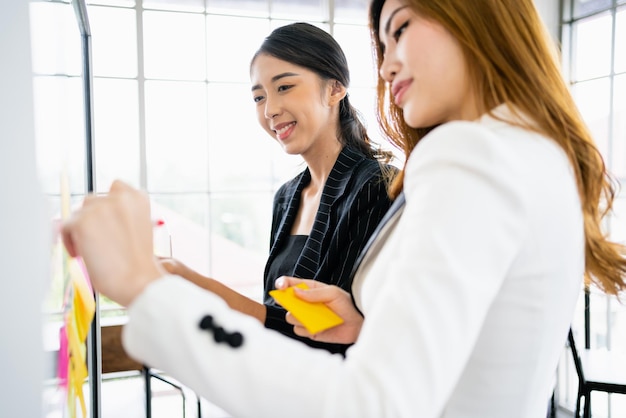 Group of successful Asian businesswomen teamwork Brainstorm meeting with colorful sticky paper notes on the glass wall for new ideas Using agile methodology for business in a tech startup office