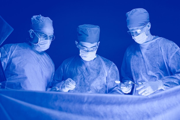 Group of surgeons at work in operating theater toned in blue medical team performing operation