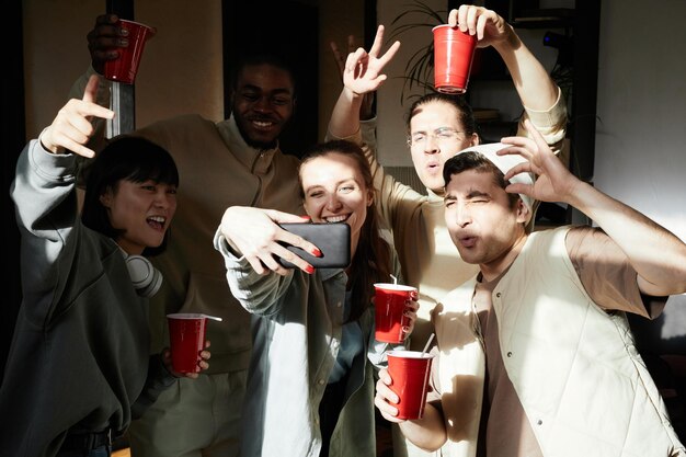 Group of teenagers with drinks posing at camera of mobile phone making selfie portrait during a party