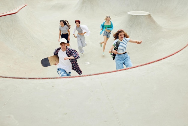 Group of teens in casual clothes boy and girls running with skate on skateboard ramp Activity and fun Concept of youth culture sport dynamic extreme hobby action and motions friendship