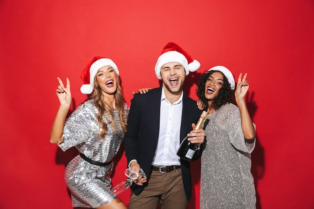 Group of three cheerful smartly dressed friends standing isolated over red space, celebrating New Year