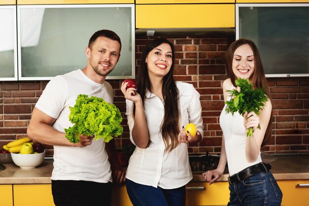 Group of three friends preparing food for dinner, posing in the kitchen holding vegetables and fruits. Promoting healthy eating. Friends are preparing a salad