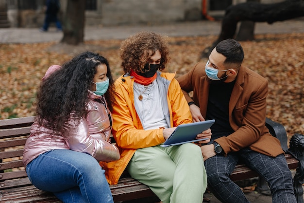 Group of three multicultural people in medical masks sitting on bench and using digital tablet. Young male and female friends relaxing at autumn park.