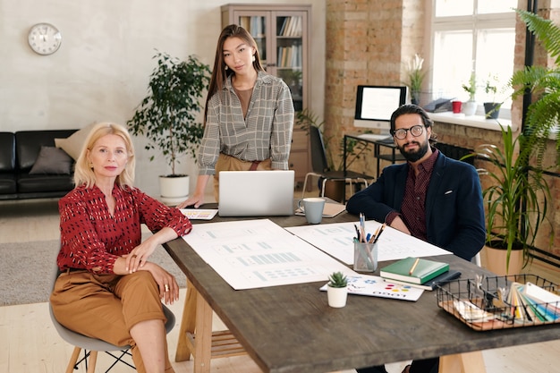 Group of two elegant females and young bearded businessman looking at you while having working meeting by large wooden table in office