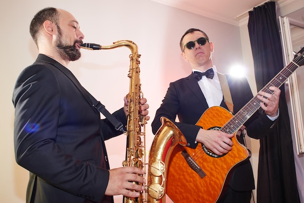 Group of two musicians, male jazz band, guitarist and saxophonist in classical costumes improvise on musical instruments in a studio stage lighting