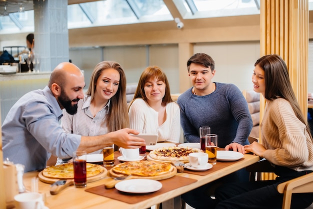 A group of young cheerful friends is sitting in a cafe talking and eating pizza