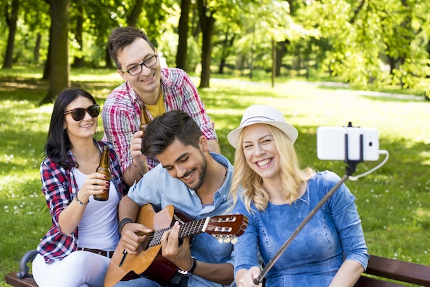 Group of young friends playing guitar and taking a selfie on a park bench