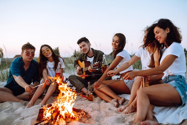 Group of young friends sitting on beach and fry sausages One man is playing guitar Camping time