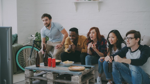 Group of young friends watching olympic games match on TV together eating snacks and drinking beer Some of them happy with their team winning but others disappointed