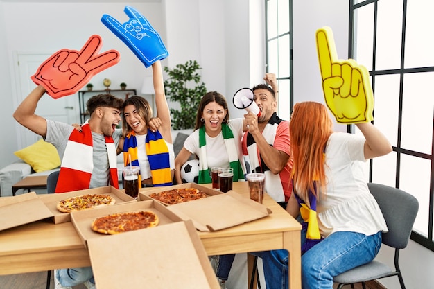 Group of young friends watching and supporting soccer match eating pizza at home.