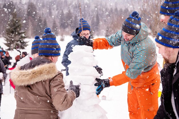 group of young happy business people having a competition in making snowmen while enjoying snowy winter day with snowflakes around them during a team building in the mountain forest