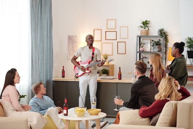 Group of young intercultural friends sitting on couches around small table with snacks and drinks and listening to singing of African guy at party