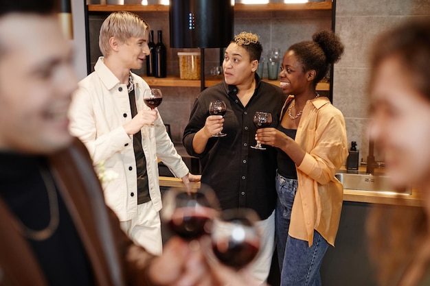 Group of young intercultural friends with glasses of red wine chatting to one another in the kitchen