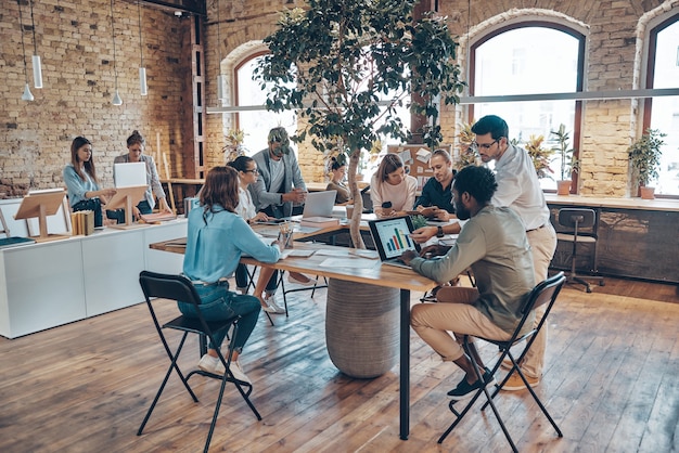 Group of young modern people in smart casual wear communicating and using modern technologies while working in the office