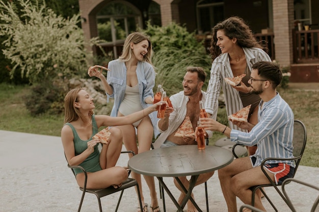 Group of young people cheering with cider and eating pizza by the pool in the garden