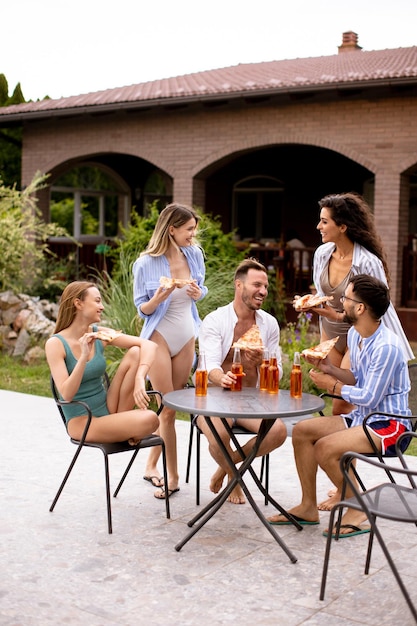 Group of young people cheering with cider and eating pizza by the pool in the garden